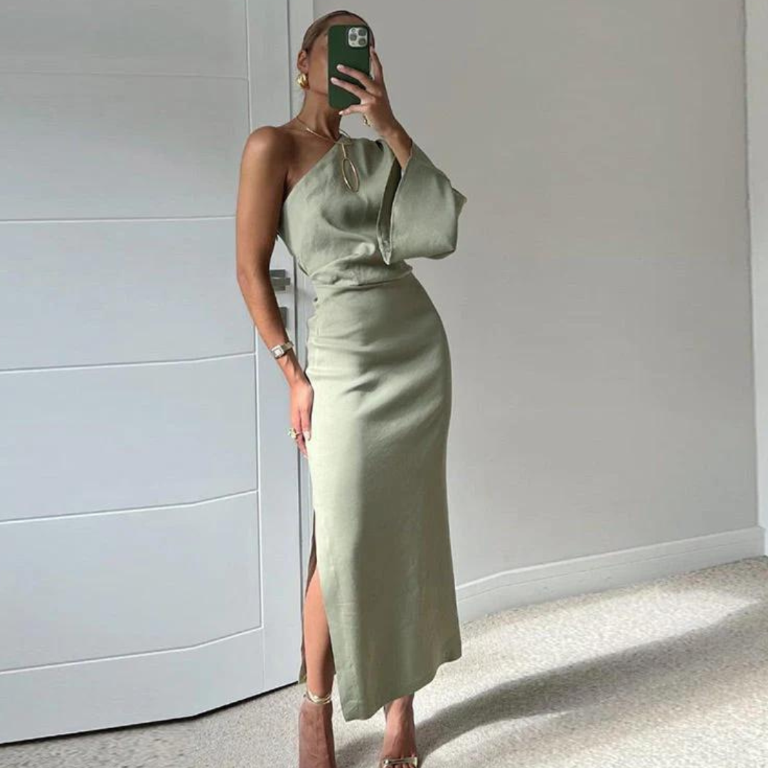 Frau in elegantem, einarmigem, olivgrünem Kleid posiert vor weißer Wand, hält Smartphone. Modetrend, Abendmode, stilvolles Outfit.