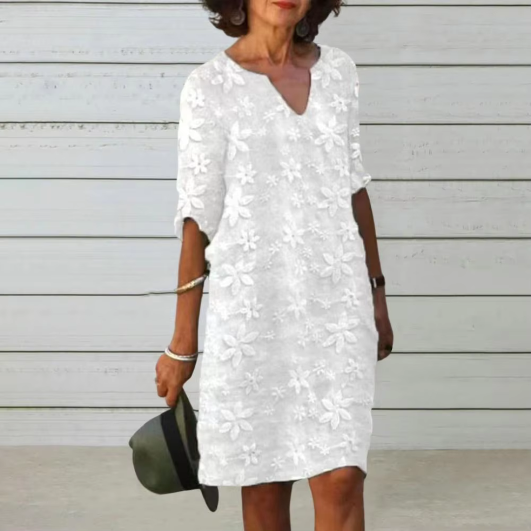 Ältere Frau in weißem, geblümtem Sommerkleid vor Holzwand, hält Hut. Modetrend, Sommermode, Damenbekleidung, Freizeitlook.