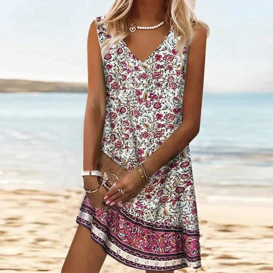 Blonde Frau in geblümtem Sommerkleid am Strand, mit Schmuck und Armbändern, vor blauem Meer und hellem Himmel. Perfekt für Strandmode und Sommerstil.