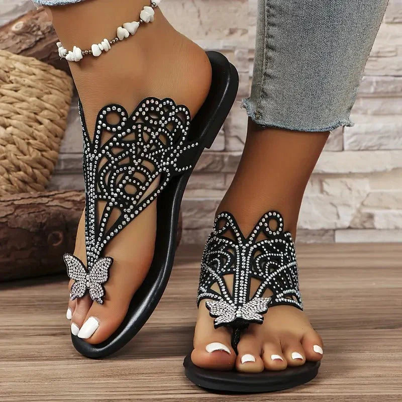 Schwarze Sandalen mit Strassverzierung und Schmetterlingsdesign, getragen von einer Person mit Jeans. Elegante Sommerschuhe für Damen.