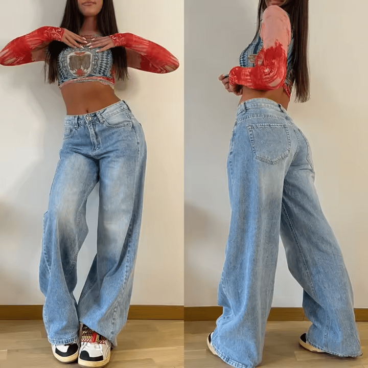 Frau in weitem Jeans und bauchfreiem Top mit langen Ärmeln, posiert vor weißer Wand. Modetrend, lässiger Stil, Streetwear-Look.