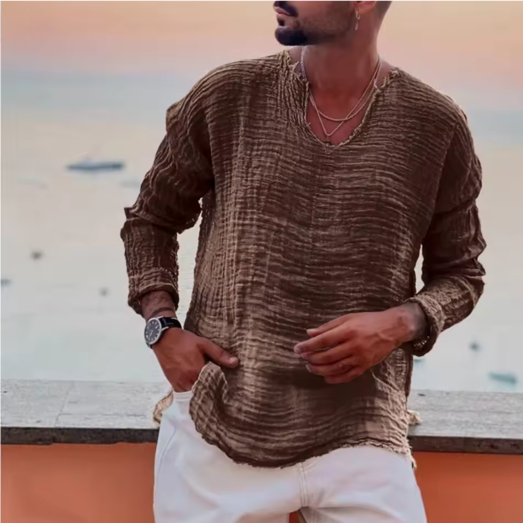 Mann in braunem Leinenhemd und weißer Hose steht vor Meereshintergrund bei Sonnenuntergang. Stilvolle Sommermode, Herrenbekleidung, Freizeitlook.