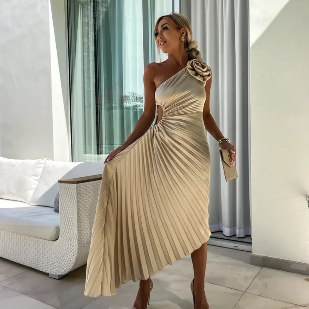 Frau in elegantem, einseitigem, beigem Abendkleid mit Plissee-Design, steht in modernem Innenraum. Perfekt für Mode, Abendmode, Stil.