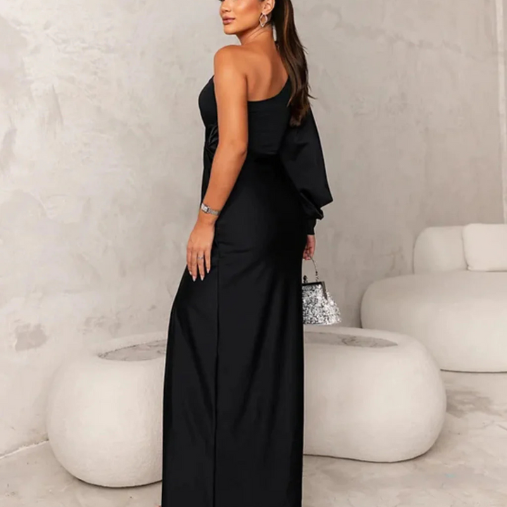 Frau in elegantem, schwarzem Abendkleid mit asymmetrischem Design, hält eine glitzernde Handtasche, steht in modernem, minimalistischem Raum.