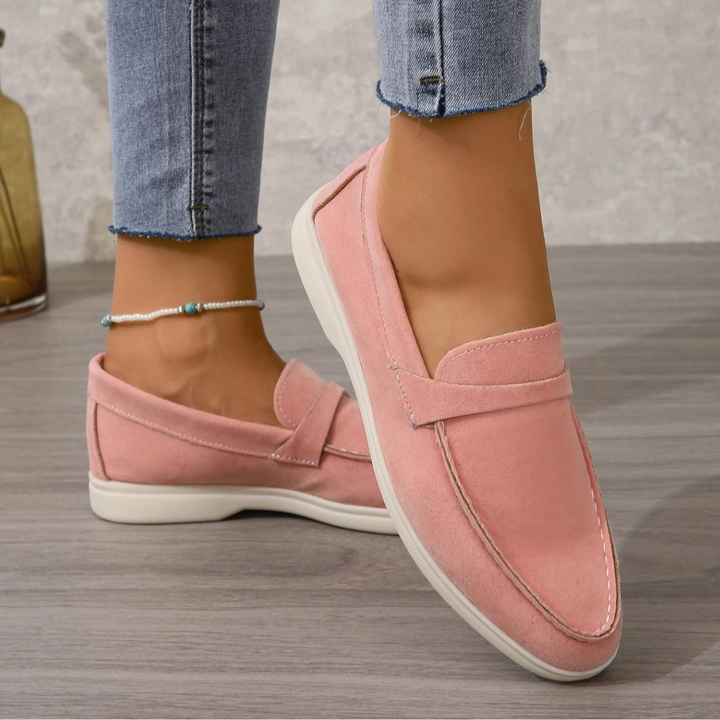 Damenfüße in rosa Mokassins auf Holzboden, kombiniert mit blauen Jeans. Modische, bequeme Schuhe für Freizeit und Alltag.