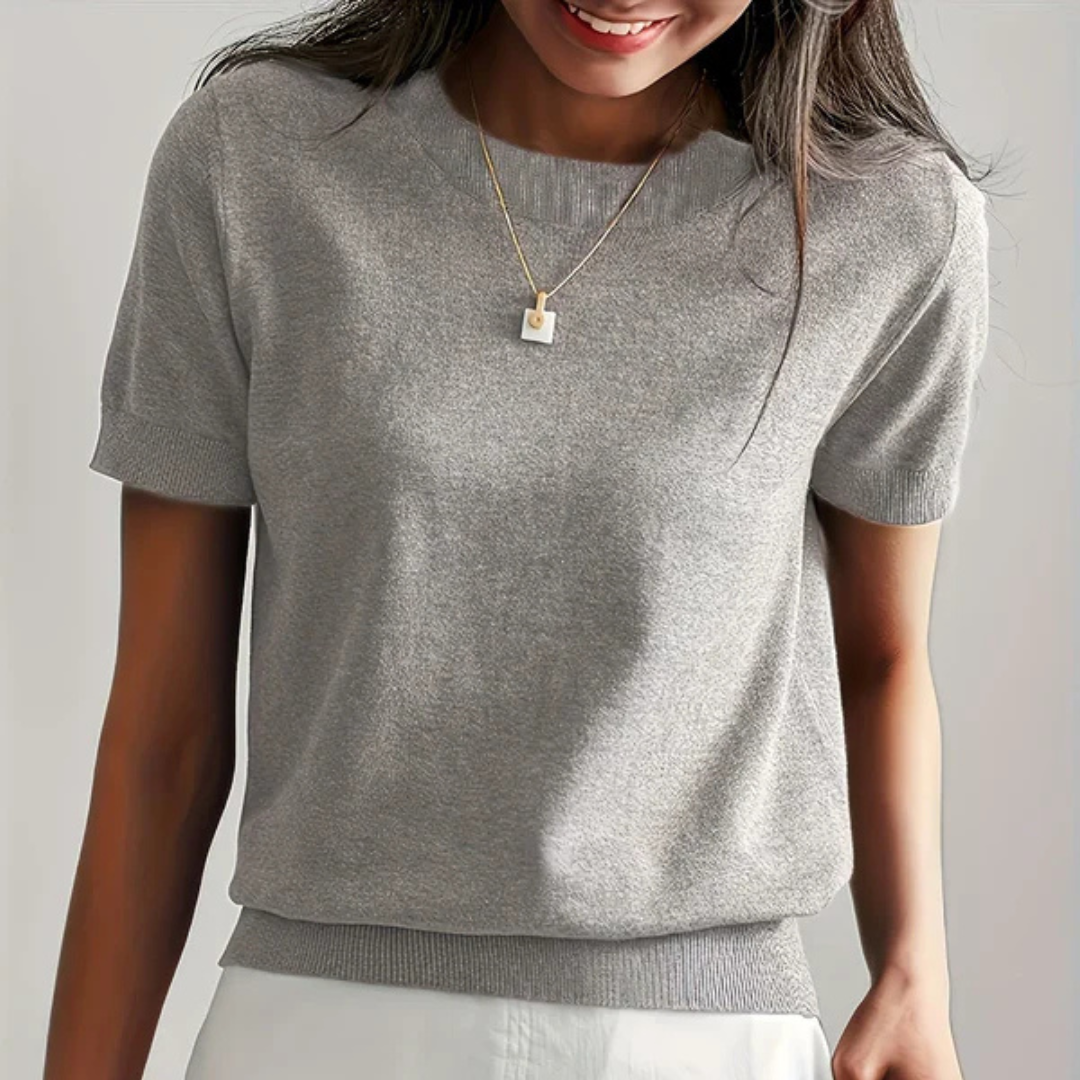 Frau trägt graues Kurzarmshirt, kombiniert mit weißem Rock und goldener Halskette. Modetrend, Damenbekleidung, eleganter Freizeitlook.
