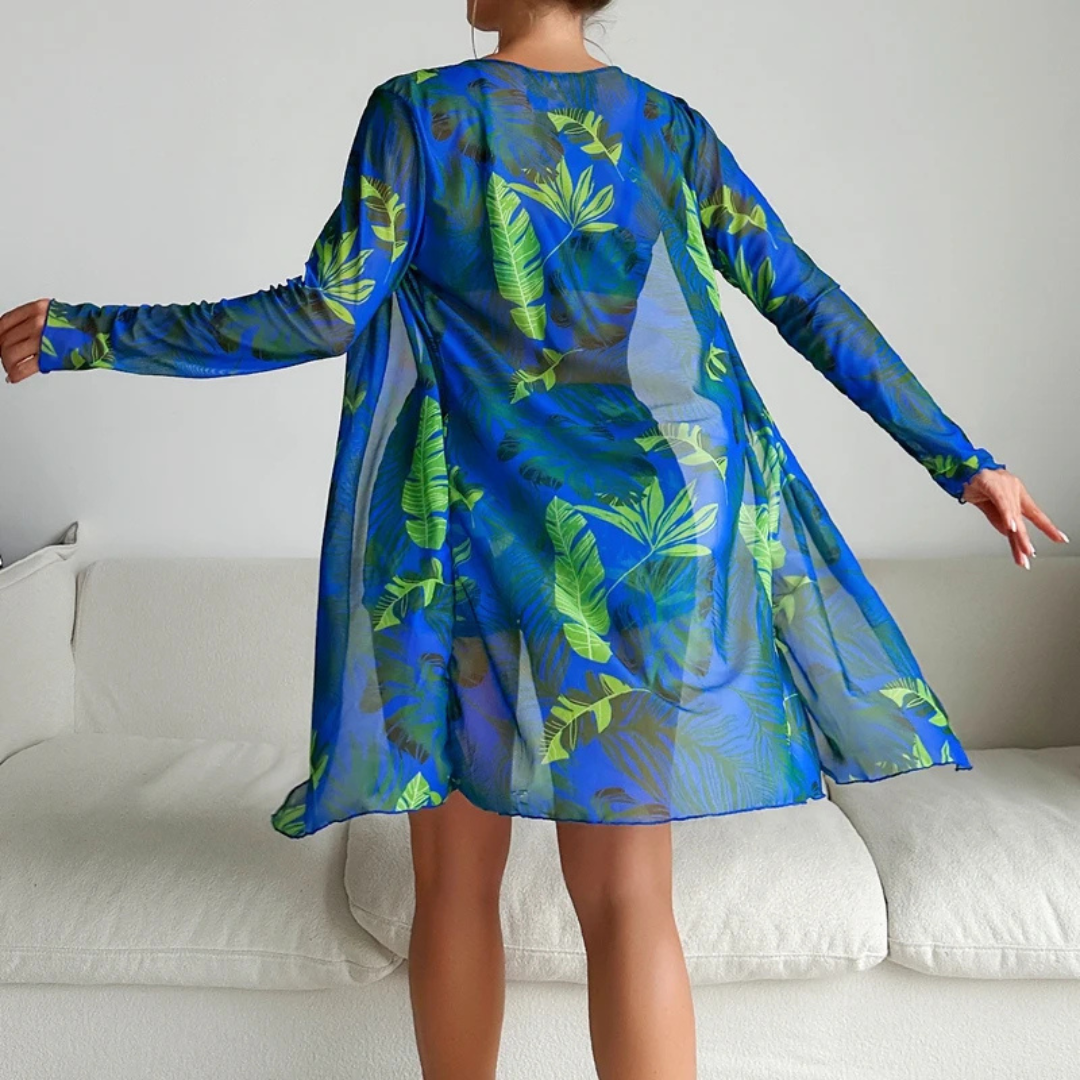 Frau in blauem, transparentem Strandkleid mit tropischem Blattmuster, steht vor weißem Sofa. Sommermode, Strandbekleidung, tropisches Design.