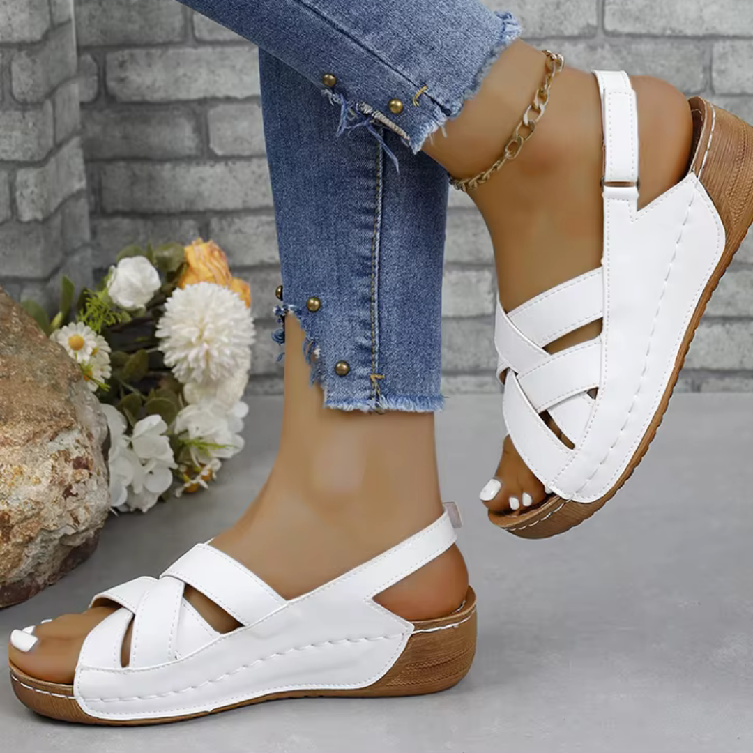 Weiße Sandalen mit Keilabsatz an Füßen in zerrissenen Jeans, vor einer Steinmauer mit Blumen. Modische Sommerschuhe, bequem und stilvoll.