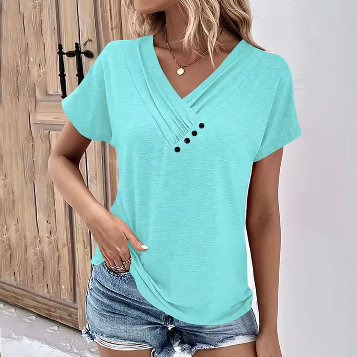 Frau in türkisfarbenem V-Ausschnitt-T-Shirt mit Knöpfen, kombiniert mit Jeansshorts, steht vor Holztür. Sommermode, lässiger Stil.