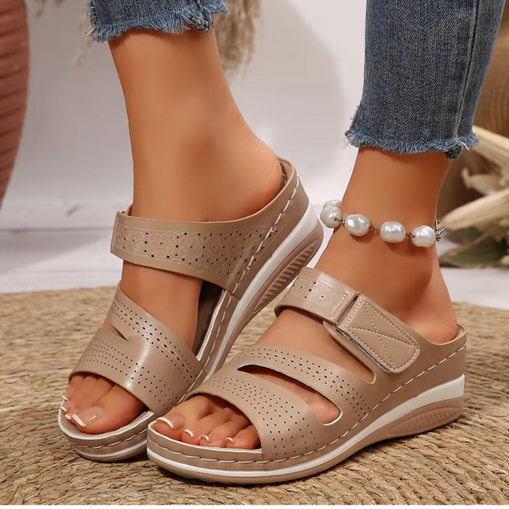 Beige Damen-Sandalen mit Keilabsatz, perforiertem Design und Klettverschluss, getragen mit Jeans und Perlenknöchelband auf Teppich.