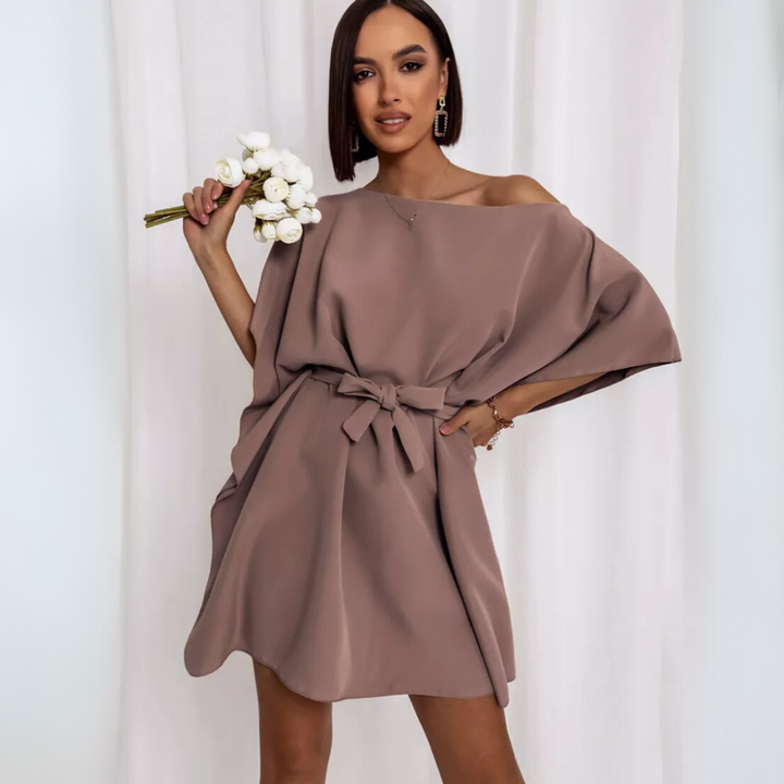 Frau in elegantem, schulterfreiem, taupefarbenem Kleid mit Schleife, hält weißen Blumenstrauß, vor weißem Hintergrund. Mode, Damenkleidung, Blumen.