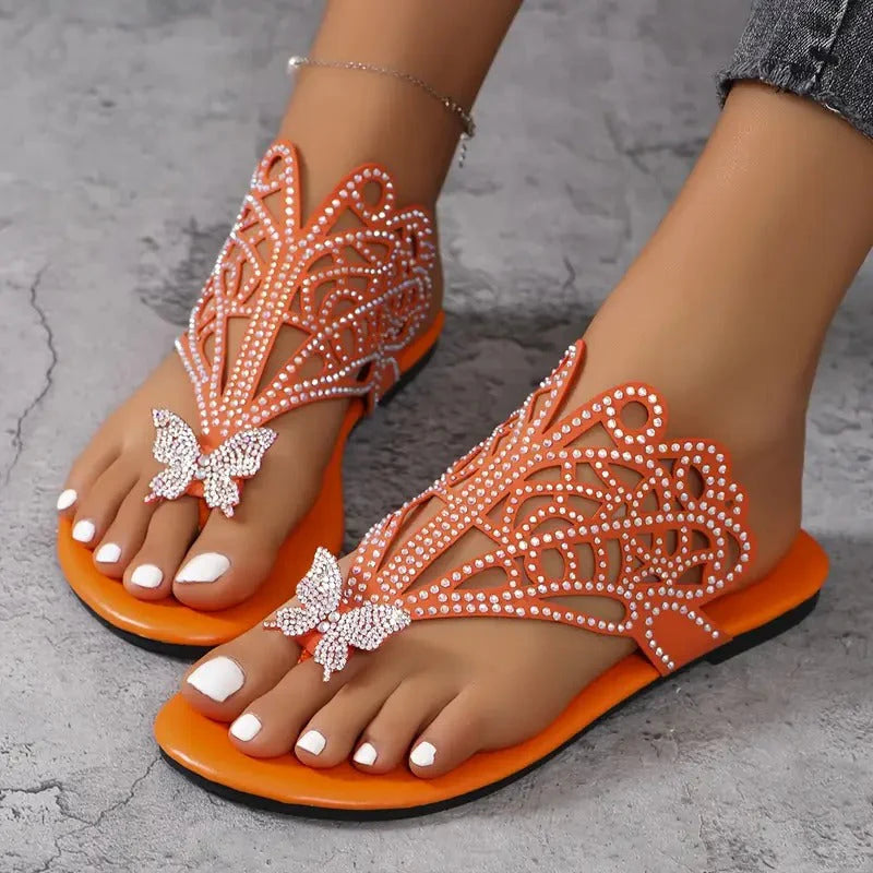 Orange Sandalen mit Schmetterlingsdesign und Strasssteinen auf grauem Hintergrund, getragen von einer Person mit weiß lackierten Zehennägeln.