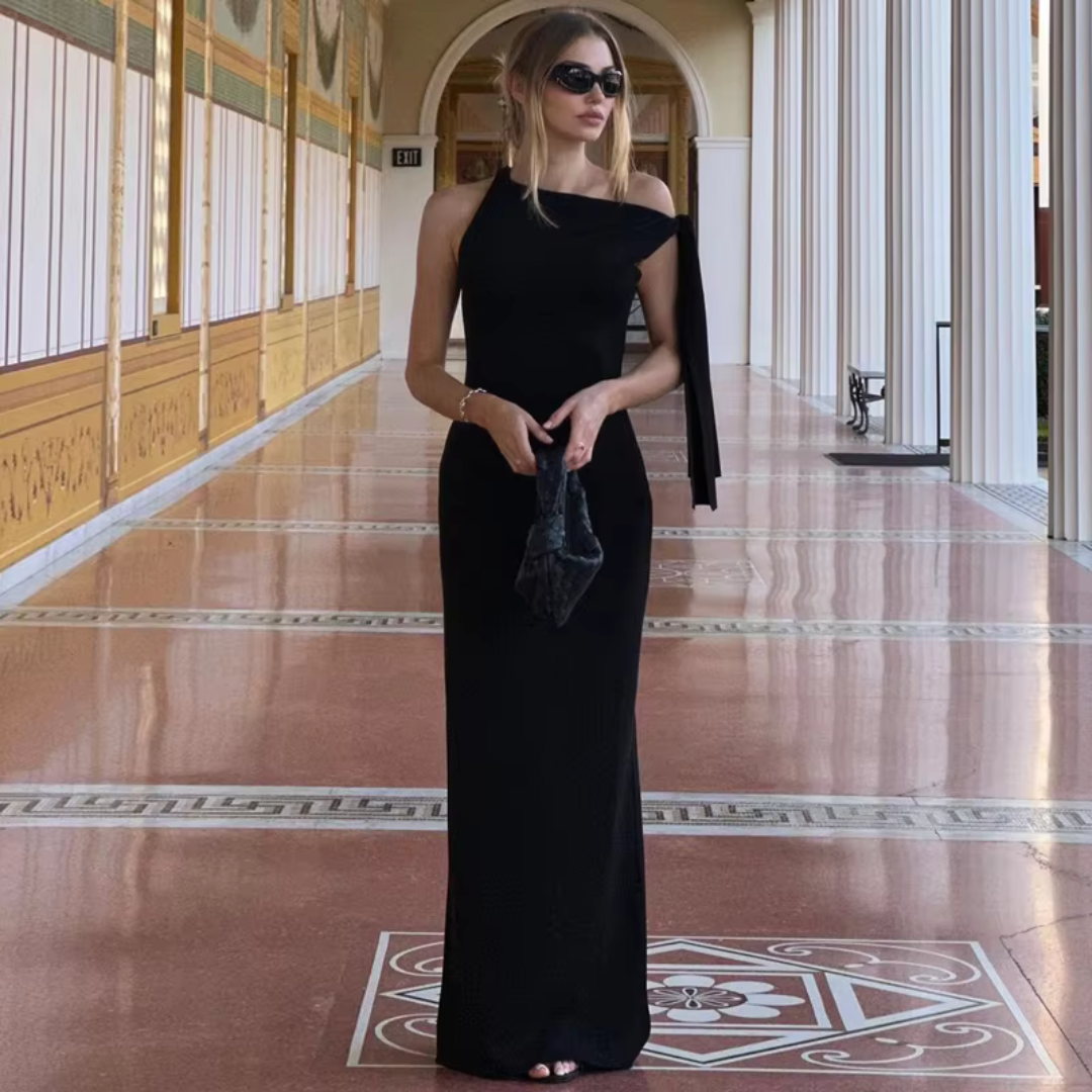 Frau in elegantem schwarzen Abendkleid mit asymmetrischem Ausschnitt, Sonnenbrille und Clutch, steht in einem luxuriösen, säulengeschmückten Flur.