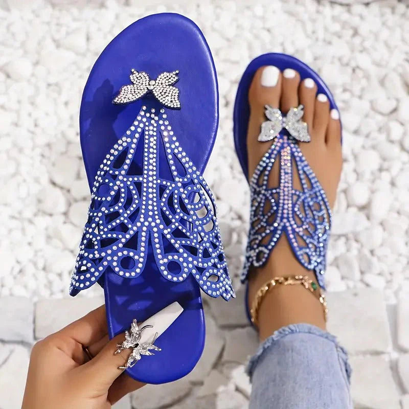 Blaue Sandalen mit Strasssteinen und Schmetterlingsdesign, getragen auf weißen Kieselsteinen. Elegante Sommerschuhe, modisch und stilvoll.