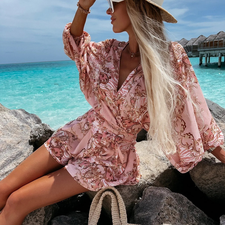 Blonde Frau in rosa Sommerkleid und Strohhut sitzt auf Felsen am türkisblauen Meer, luxuriöse Strandkulisse, tropischer Urlaub, Mode.