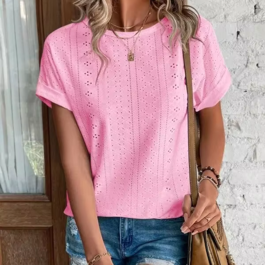 Frau in rosa Lochstickerei-T-Shirt und Jeansshorts vor weißer Backsteinwand, trägt braune Umhängetasche. Sommermode, Freizeitlook.