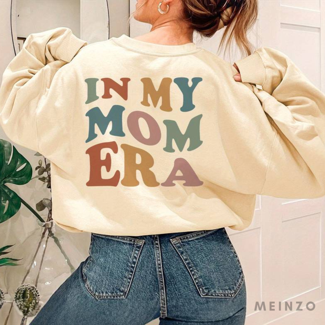 Frau trägt beigen Pullover mit buntem Schriftzug "In My Mom Era", kombiniert mit blauer Jeans. Modetrend, lässiger Stil, Retro-Design.