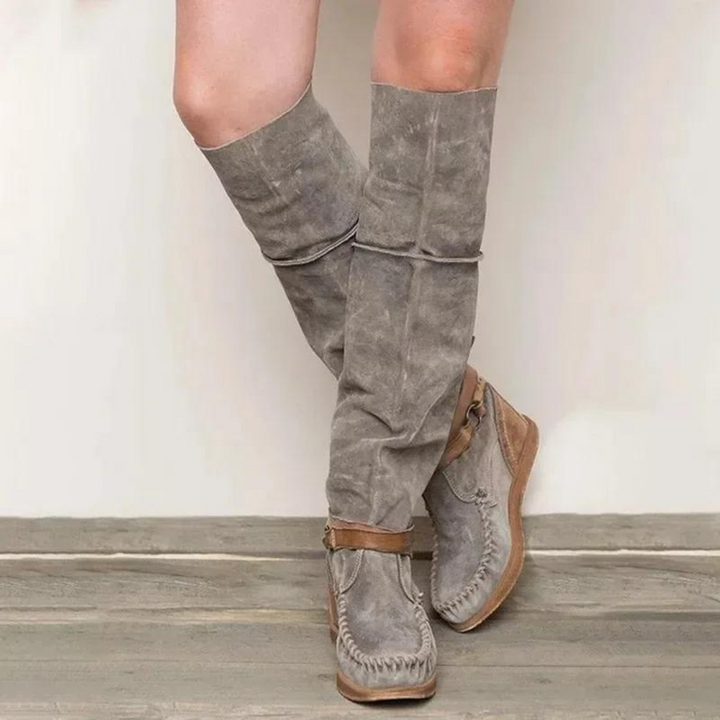 Graue Wildlederstiefel mit braunen Ledersohlen auf Holzboden. Modische Damenstiefel, knielang, ideal für Herbstmode und Outdoor-Aktivitäten.