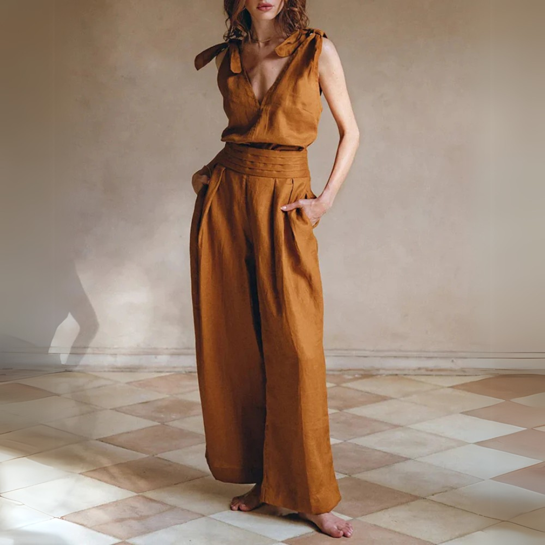 Frau in braunem Leinen-Jumpsuit mit weitem Bein und V-Ausschnitt, steht barfuß auf Fliesenboden. Elegante, nachhaltige Damenmode.