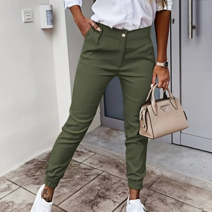 Frau in grüner Hose und weißem Hemd, hält beige Handtasche, trägt weiße Sneaker. Modischer Streetstyle-Look, ideal für urbane Outfits.