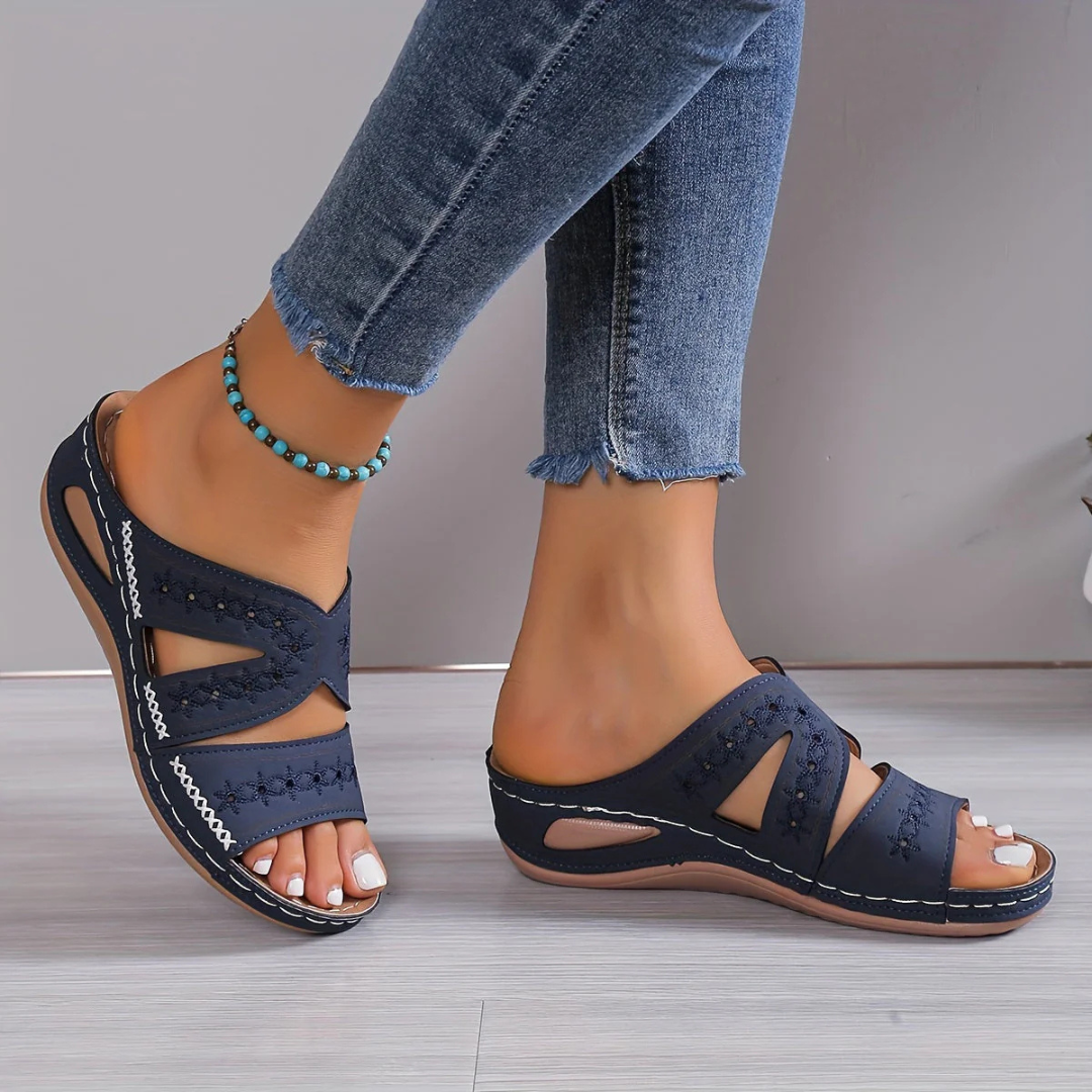 Damenfüße in blauen Sandalen mit Cut-Out-Design, kombiniert mit Jeans und einem türkisfarbenen Fußkettchen. Sommermode, bequeme Schuhe, Freizeitlook.