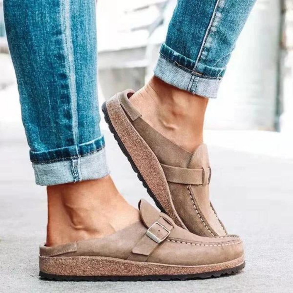 Beige Damen-Clogs aus Wildleder mit Schnalle, kombiniert mit hochgekrempelten blauen Jeans. Bequeme, modische Schuhe für den Alltag.