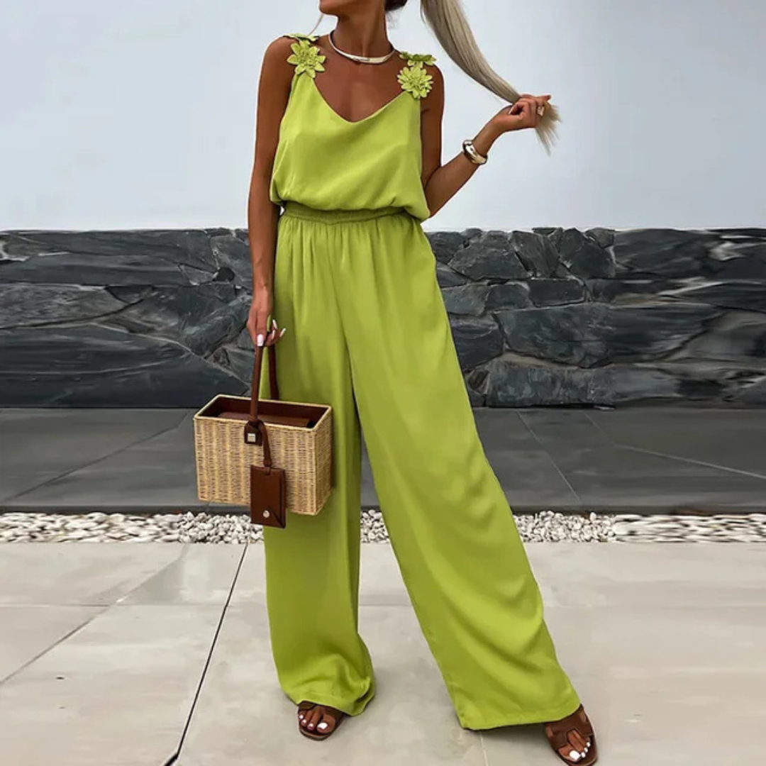 Frau in grünem Jumpsuit mit Blumenapplikationen, hält eine Strohtasche. Sommermode, eleganter Look, Outdoor-Hintergrund.