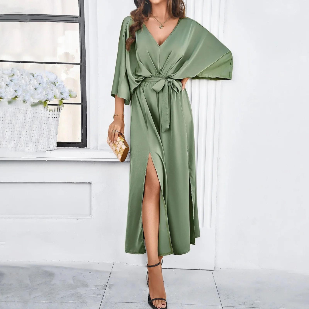 Frau in elegantem, grünem Satinkleid mit V-Ausschnitt und Schlitz, hält goldene Clutch, steht vor Fenster mit weißen Blumen. Abendmode, Damenkleider.