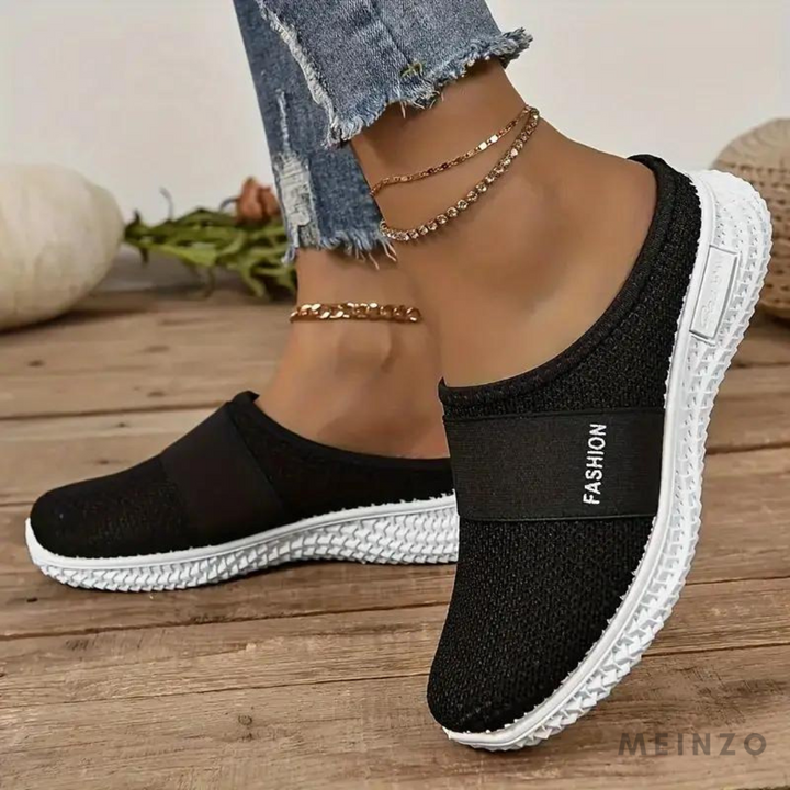 Schwarze Damen-Sneaker mit weißer Sohle, getragen mit blauen Jeans und Fußkettchen, auf Holzboden. Modische Freizeitschuhe für Damen.