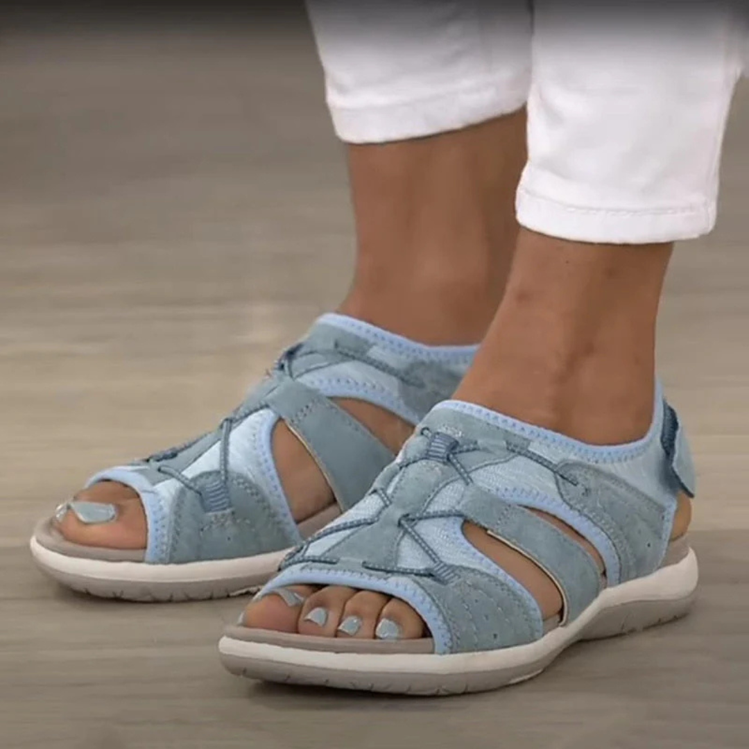Blaue Damen-Sandalen mit Klettverschluss, getragen mit weißen Hosen. Bequeme, atmungsaktive Sommerschuhe auf hellem Holzboden.