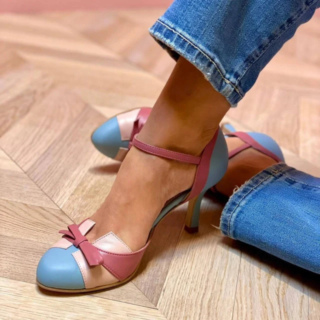 Frau trägt modische, mehrfarbige High Heels mit Schleifen-Detail auf Parkettboden. Trendige Damenschuhe, elegantes Design, Jeans.