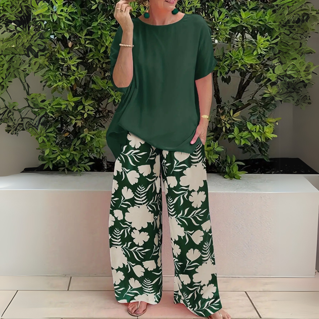 Frau in grünem Oberteil und geblümter Hose vor grüner Hecke. Sommermode, lässiger Stil, florales Muster, Outdoor-Hintergrund.