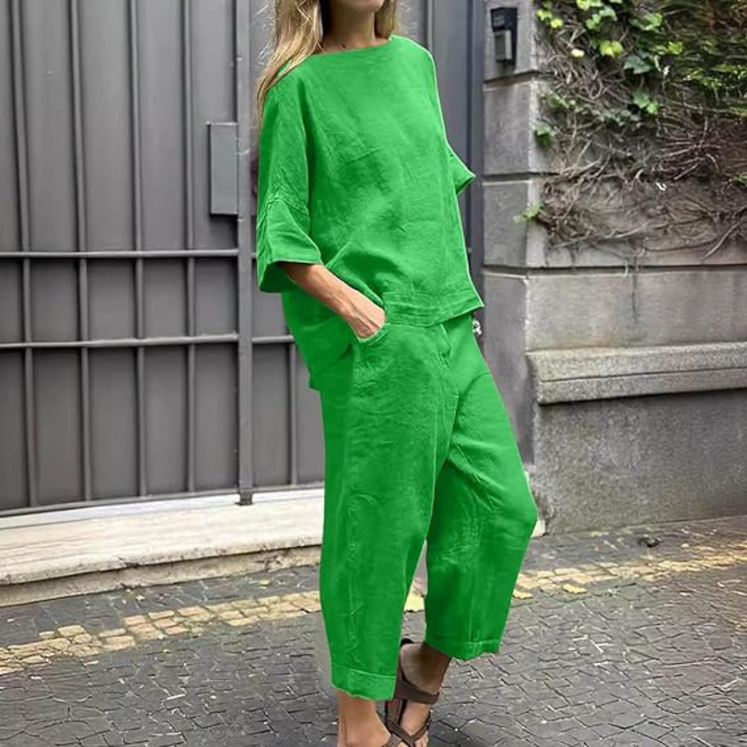 Frau in grünem Leinen-Zweiteiler, bestehend aus lockerem Oberteil und Hose, steht vor grauem Tor. Modetrend, Sommeroutfit, lässiger Stil.