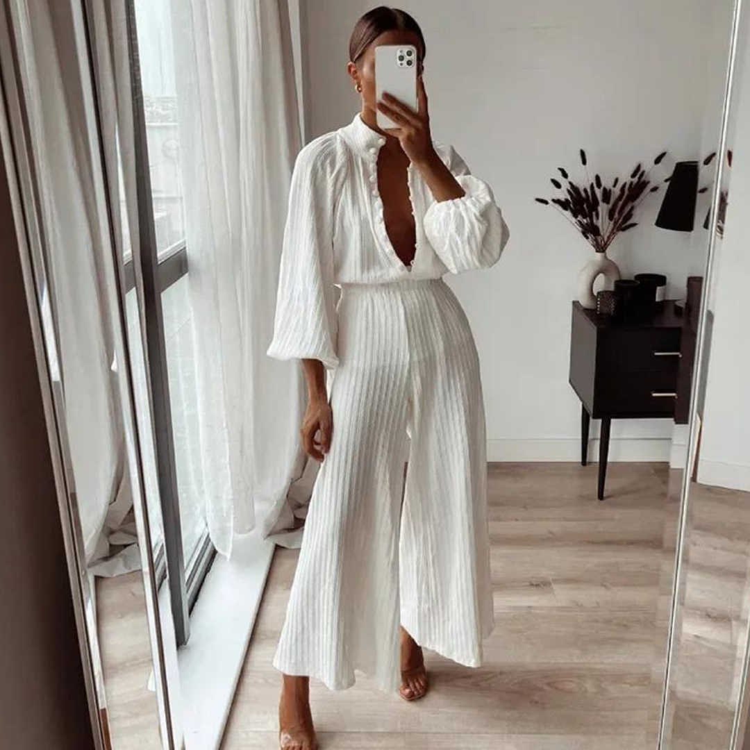 Frau in elegantem, weißem Jumpsuit mit tiefem Ausschnitt, steht vor einem Spiegel in modernem, minimalistischem Raum mit großen Fenstern und Dekoration.