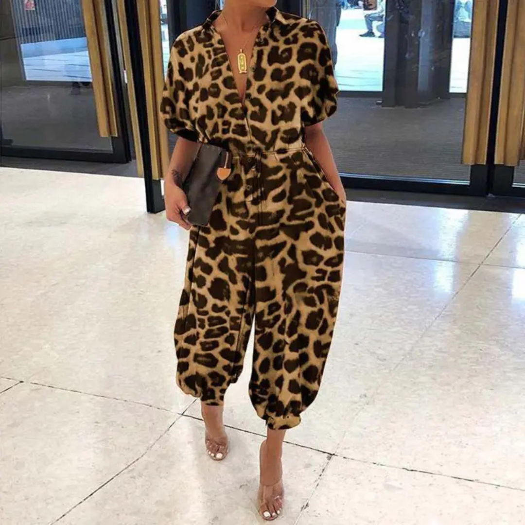 Frau in modernem Leopardenmuster-Jumpsuit, hält schwarze Clutch, steht in elegantem Innenraum. Modetrend, Damenmode, stilvolles Outfit.