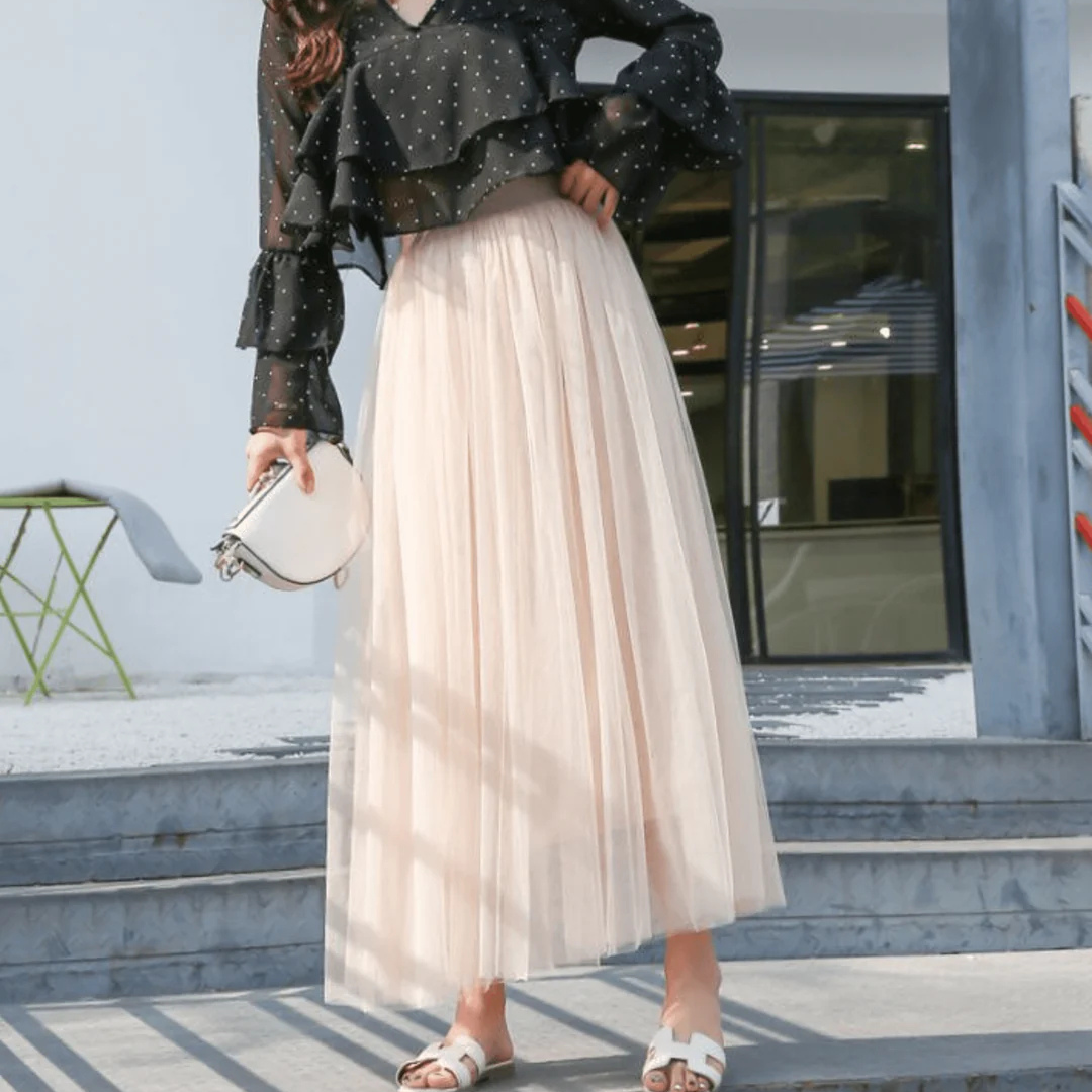 Frau in schwarzer, gepunkteter Bluse und langem, beigem Tüllrock, hält weiße Handtasche, steht auf Treppe. Elegante Mode, Sommeroutfit.