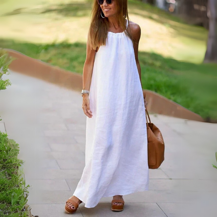 Frau in weißem Leinenkleid spaziert im Freien, trägt braune Sandalen und eine braune Tasche. Sommermode, lässiger Stil, elegantes Outfit.