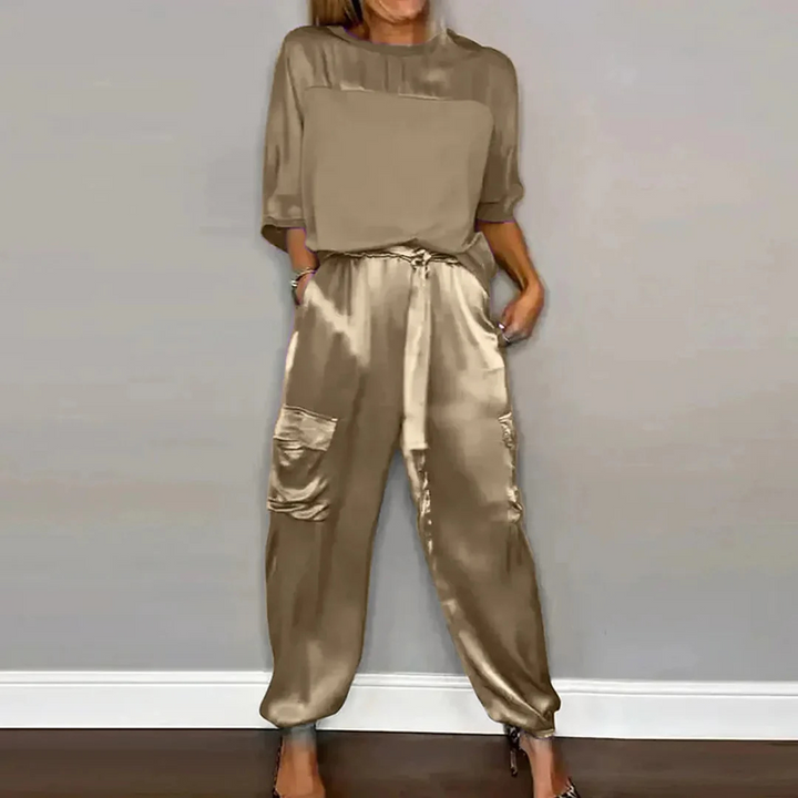 Dame in eleganter, beiger Satin-Hose mit Taschen und passendem Oberteil vor grauer Wand. Modetrend, stilvolle Damenbekleidung, Satin-Outfit.