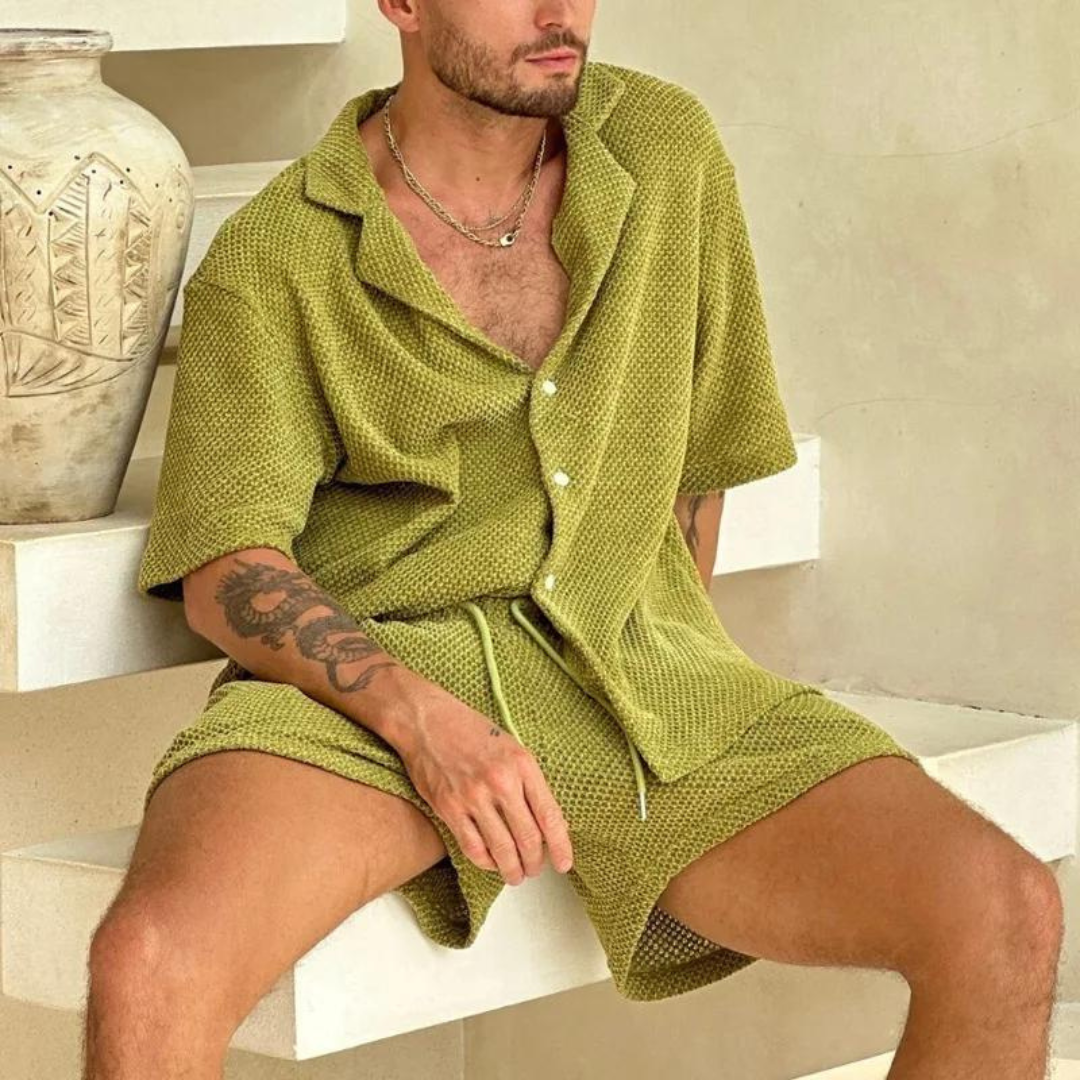 Mann in olivgrünem, strukturiertem Kurzarmhemd und Shorts sitzt auf Treppe. Sommermode, lässiger Stil, Herrenbekleidung, Tattoo am Arm.