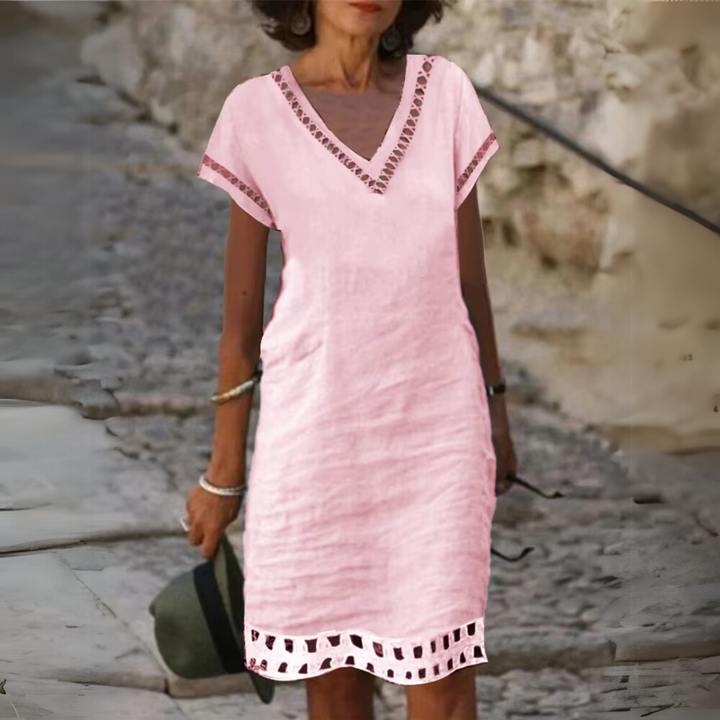 Frau in rosa Leinenkleid mit V-Ausschnitt und Spitzenbesatz, steht auf gepflastertem Weg. Sommermode, lässiger Stil, elegantes Design.