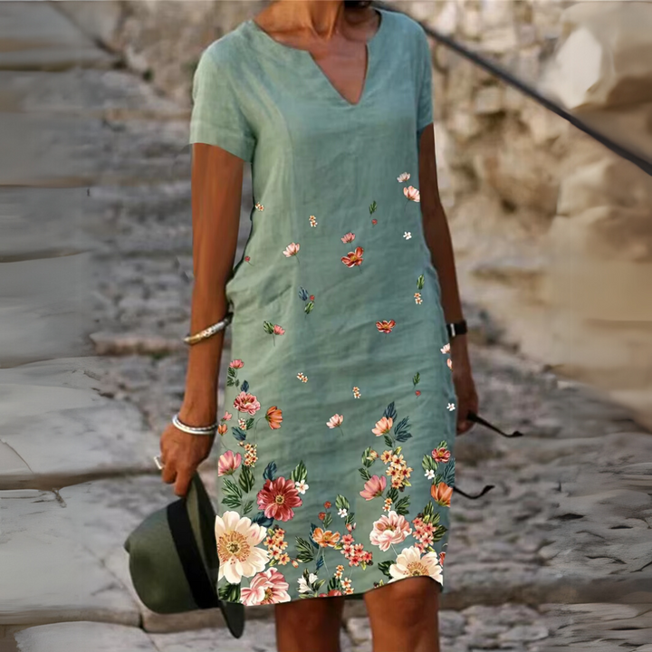Frau in grünem Sommerkleid mit Blumenmuster, hält Hut, steht auf gepflastertem Weg. Modetrend, Damenmode, Freizeitkleidung, Sommeroutfit.