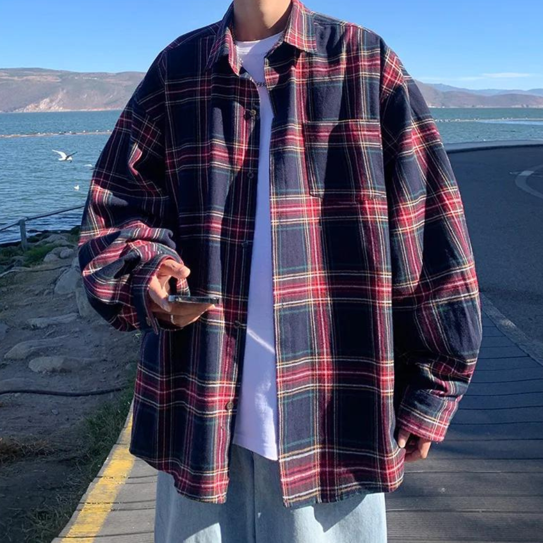 Person in übergroßem karierten Hemd am Seeufer, hält Smartphone. Modetrend, lässiger Stil, Outdoor-Hintergrund, blau-rotes Karomuster.