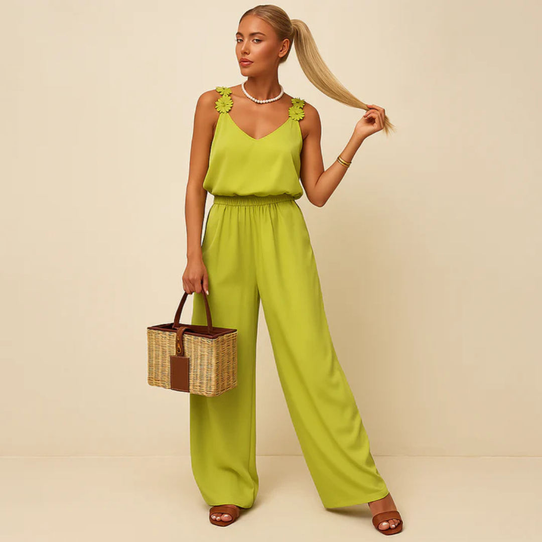 Frau in grünem Jumpsuit mit Blumenmuster-Trägern, hält ihren Pferdeschwanz. Sie trägt braune Sandalen und hält einen Korb. Elegante Sommermode.
