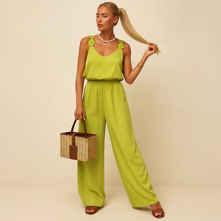 Frau in grünem Jumpsuit mit Blumenmuster-Trägern, hält ihren Pferdeschwanz. Sie trägt braune Sandalen und hält einen Korb. Elegante Sommermode.