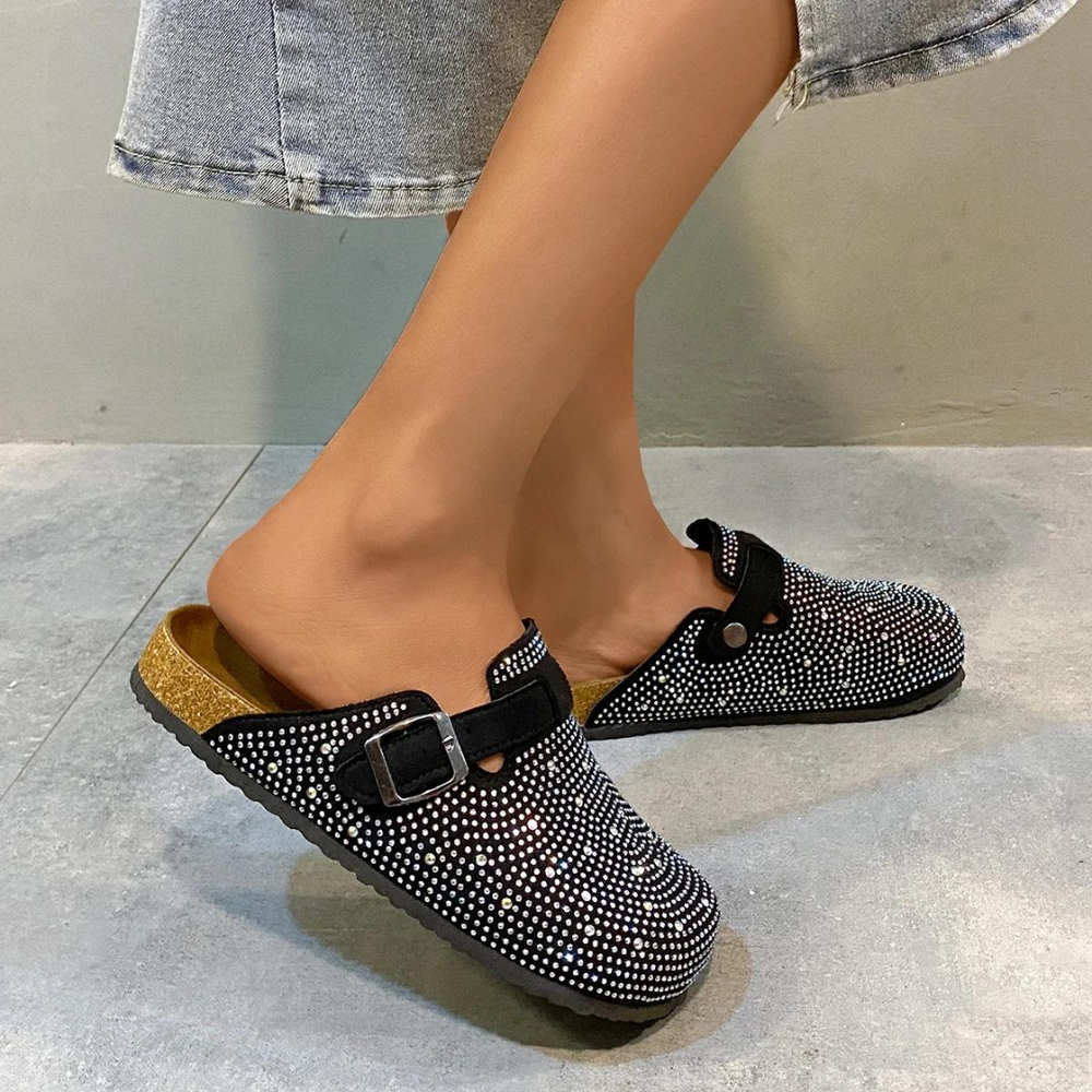 Damenfüße in glitzernden Clogs mit Strasssteinen, kombiniert mit Jeansrock. Modische, bequeme Schuhe für Alltag und Freizeit.