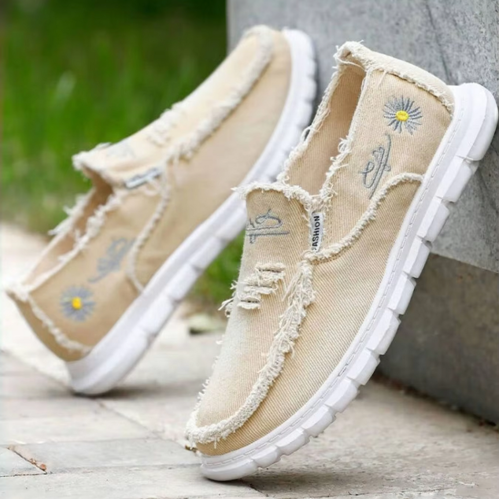 Beige Freizeitschuhe mit Gänseblümchen-Stickerei, abgenutztem Look und weißer Sohle auf Gehweg. Modische, bequeme Sommerschuhe für Damen.