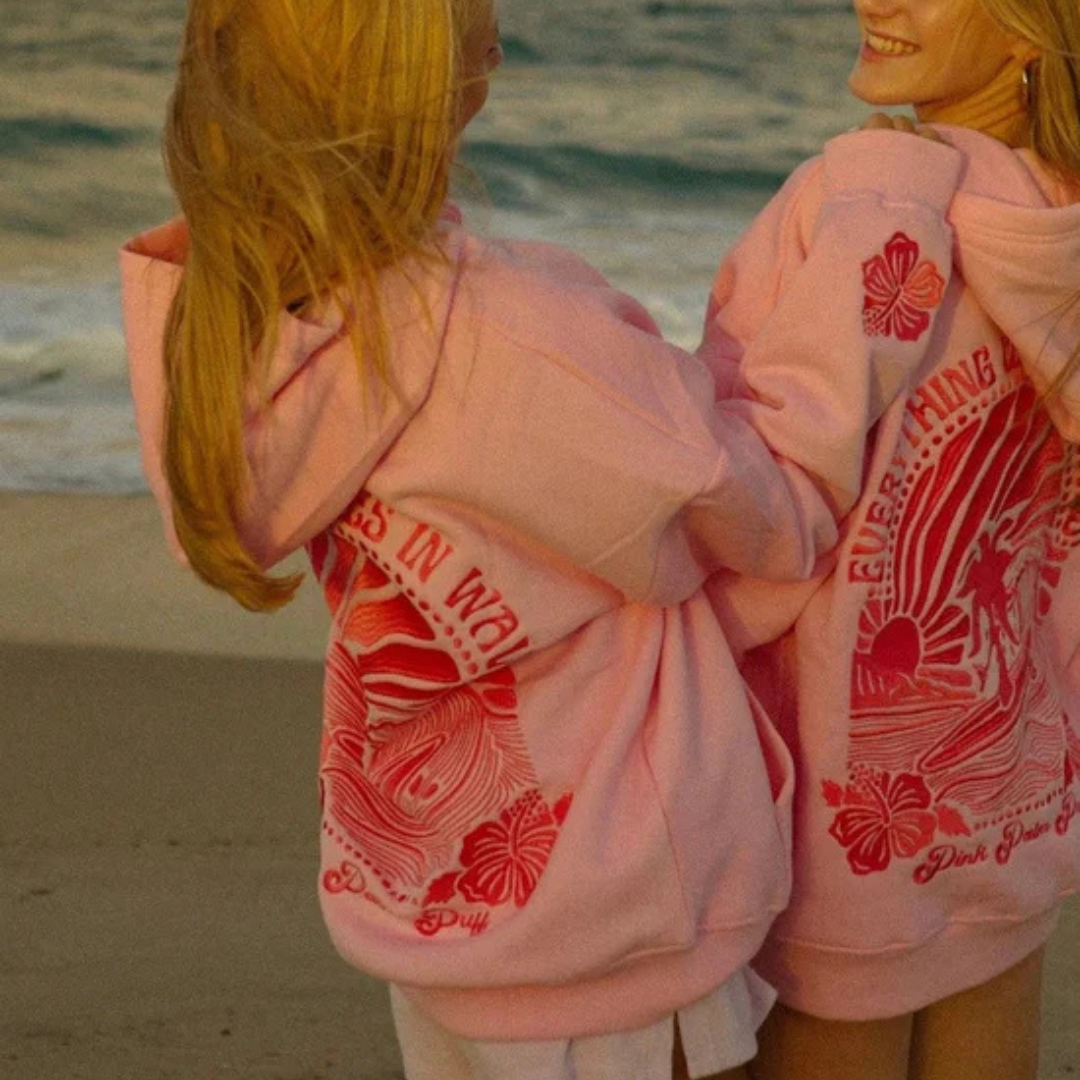 Zwei Mädchen in rosa Hoodies mit Surfmuster am Strand, lächelnd und umarmend, vor einer Meereskulisse bei Sonnenuntergang.