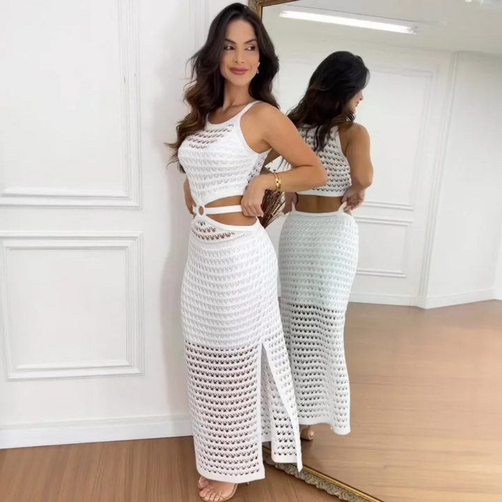 Frau in weißem, gehäkeltem Kleid mit Schlitz, posiert vor einem Spiegel in modernem Raum. Elegante Sommermode, stilvolle Damenbekleidung.