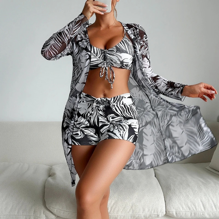 Frau in tropischem Bikini-Set mit Blätter-Print und passendem Kimono, posiert auf weißem Sofa. Sommermode, Strandoutfit, tropisches Design.
