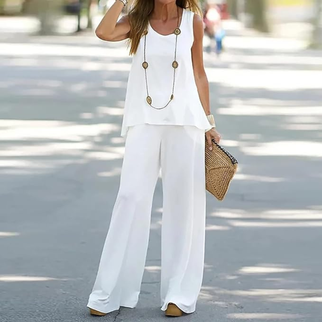 Frau in weißem Sommeroutfit mit weitem Top und Hose, trägt lange Halskette und Strohtasche, modisch und elegant auf sonniger Straße.